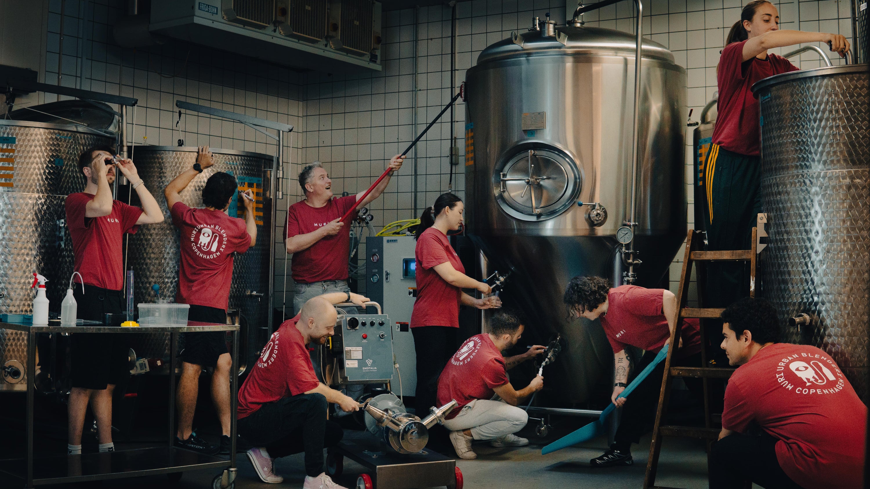 Group of people in red shirts working around large metal tanks in a brewery setting.