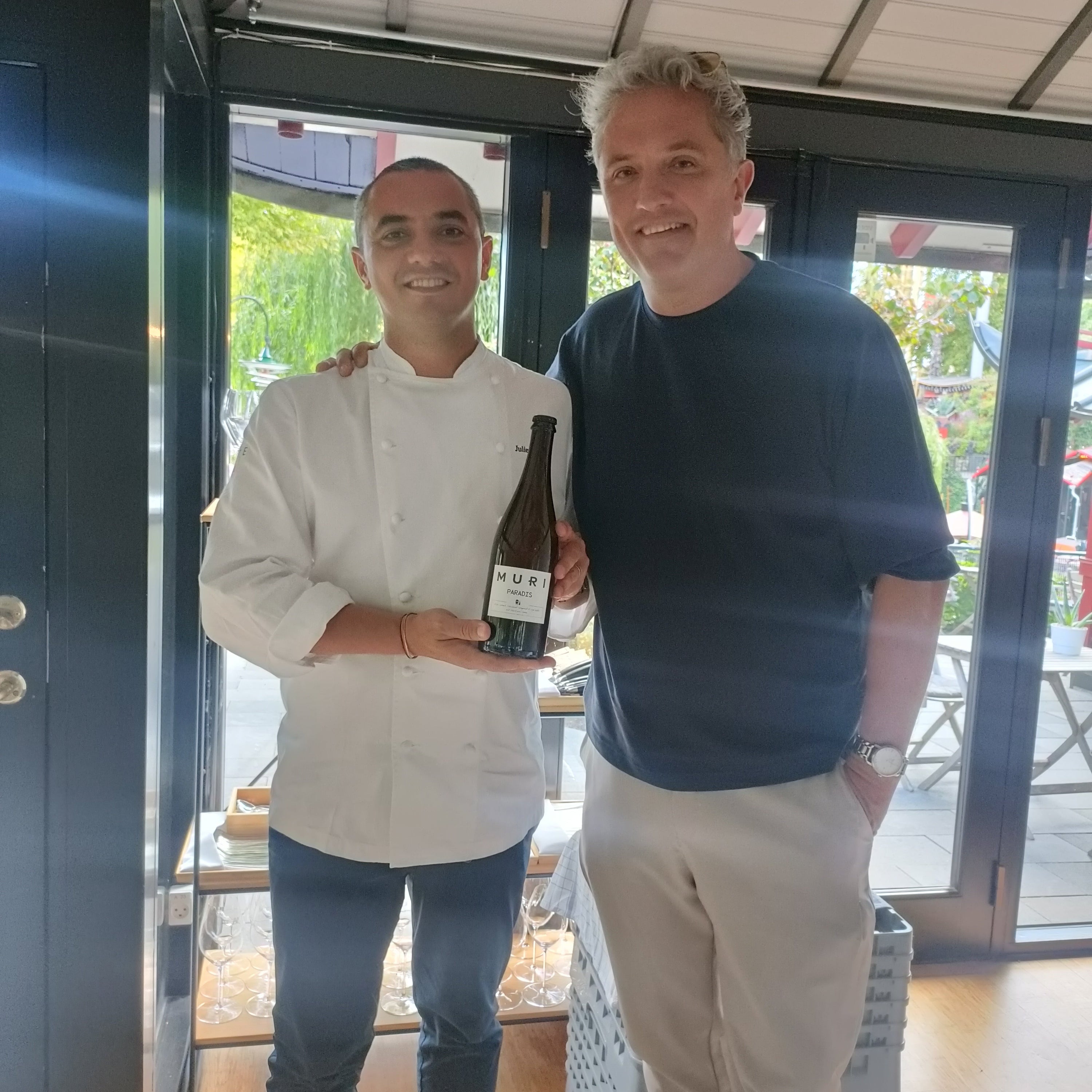 Two men standing together, one holding a bottle of Paradis, in an outdoor setting.